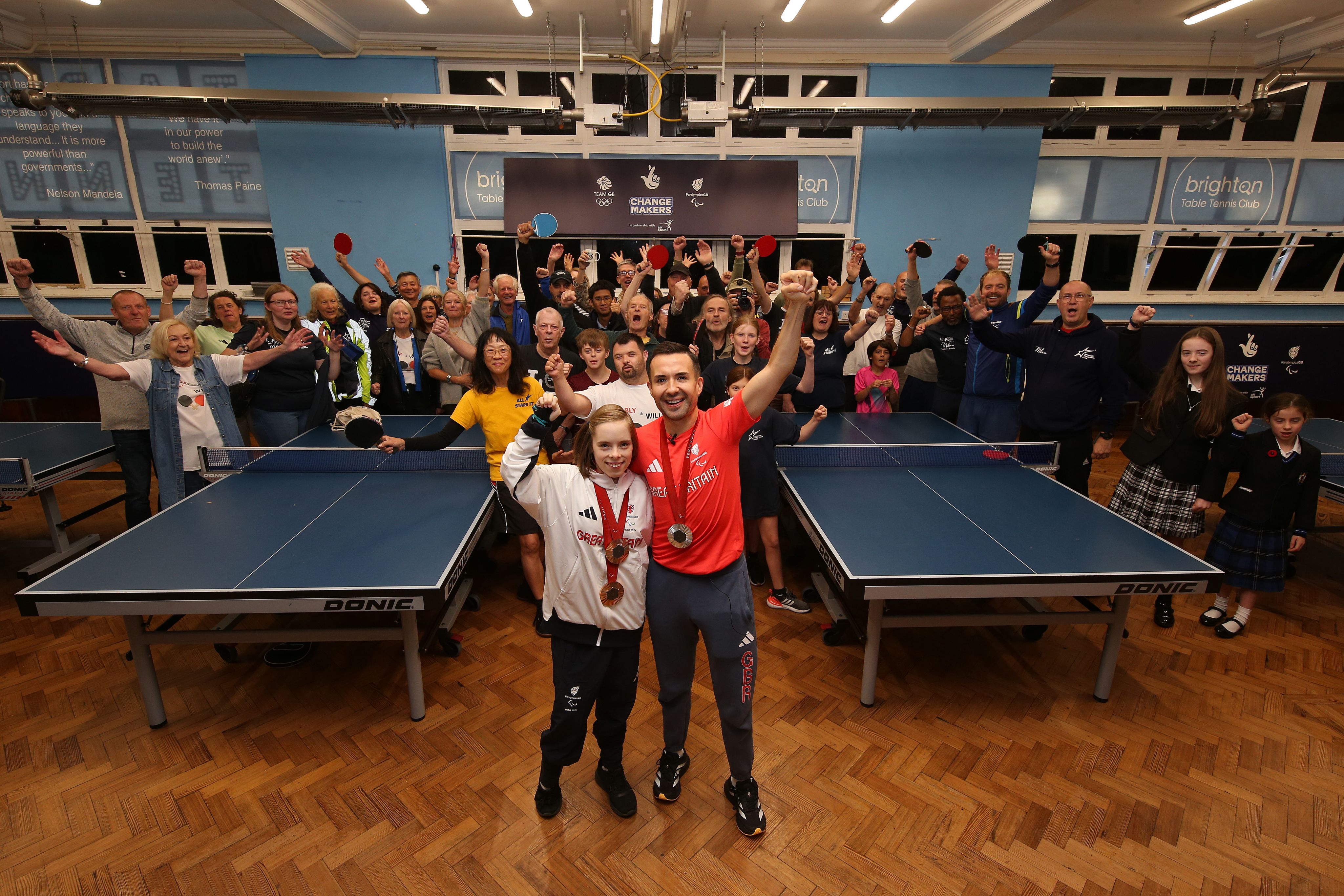 Athletes Bly Twomey and Will Bayley raise there arms in celebration in front of a group of people at Brighton Table Tennis Club.