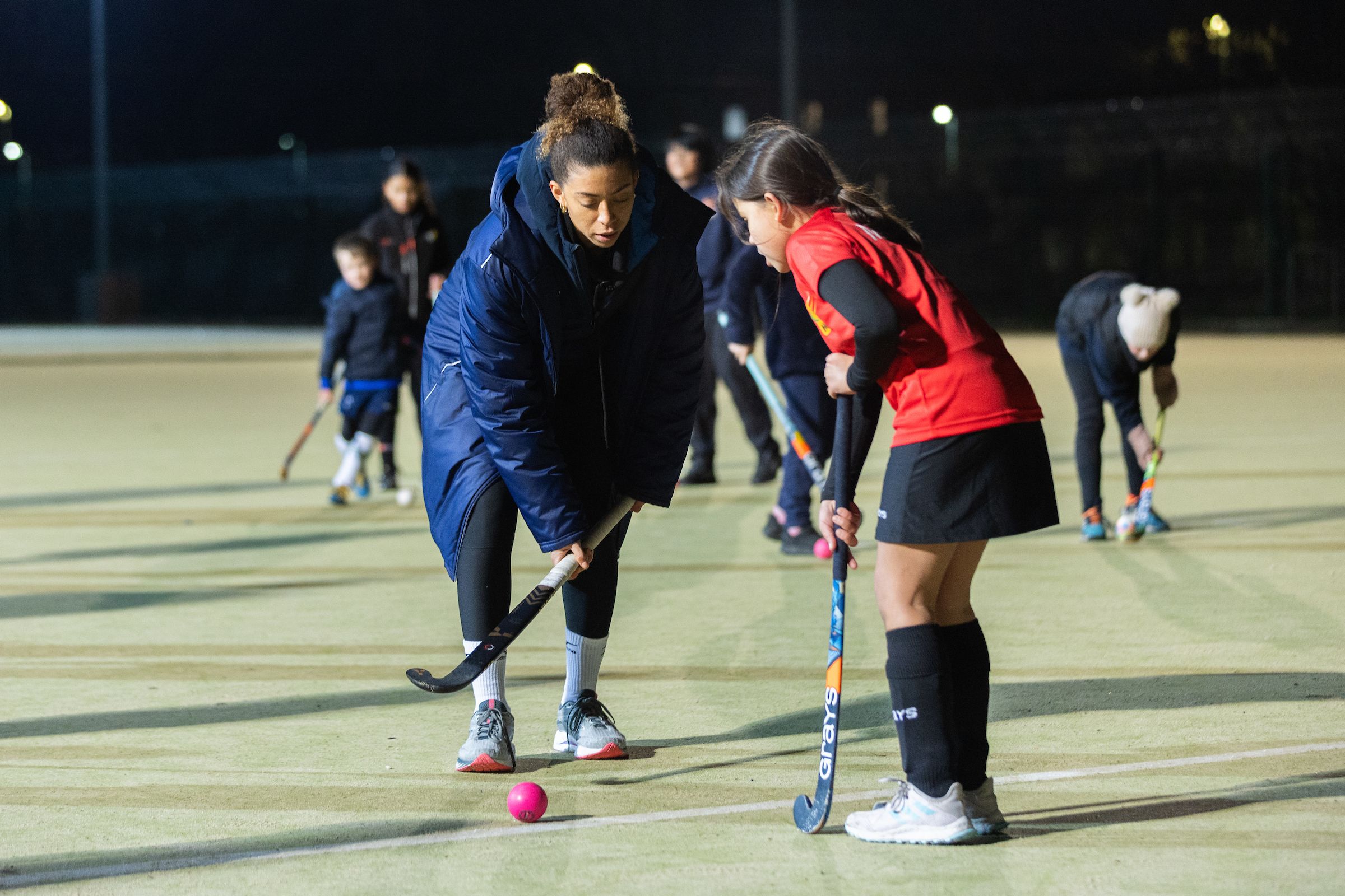Athlete Darcy Bourne coaches a young girl at hockey. Darcy is leaning over with the ball at her feet, the girl is watching as Darcy shows her what to do. They are stood on a grass pitch with other people out of focus in the background being coached at hockey.