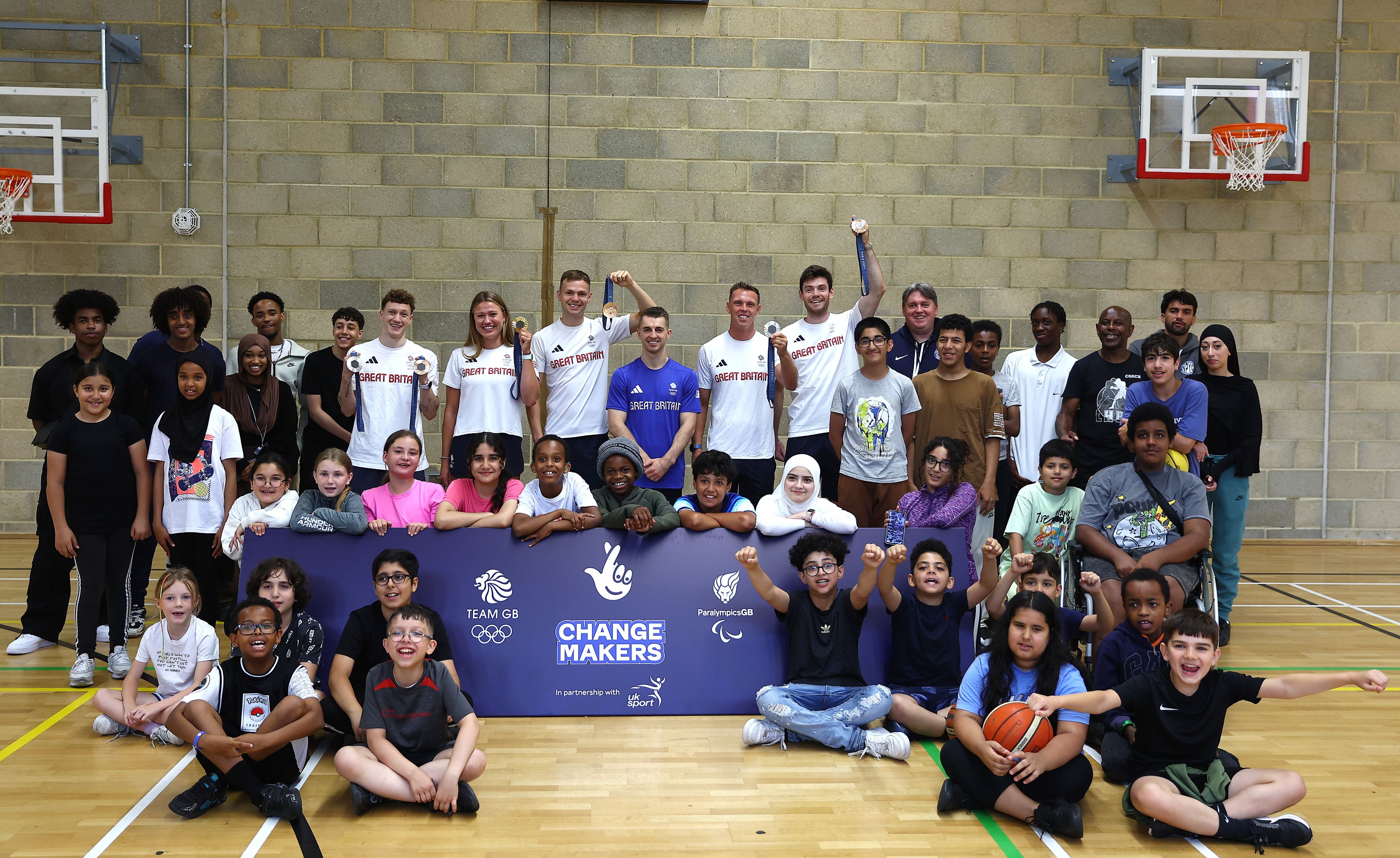 Rowers Lola Anderson and Freddie Davidson, boxer Lewis Richardson, diver Noah Williams, canoeist Joe Clarke MBE, and gymnast Max Whitlock OBE stand in a line alongside children and a banner reading ChangeMakers at a youth club to promote ChangeMakers.