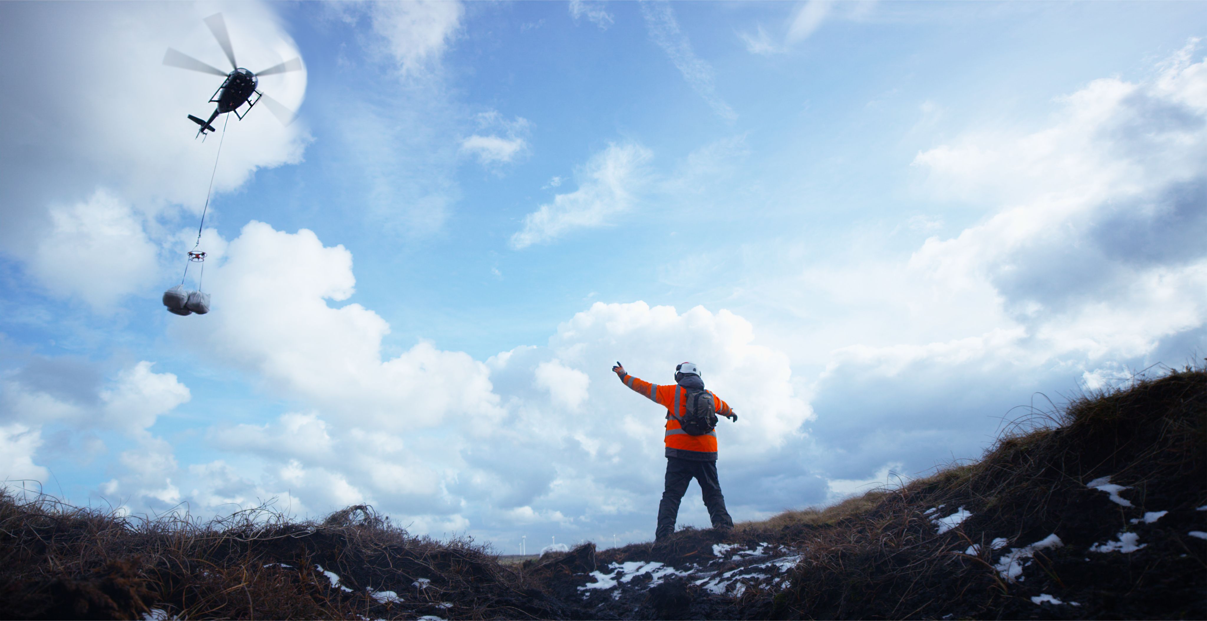 A person waves down packages that are being delivered by a helicopter on top of a mountain.