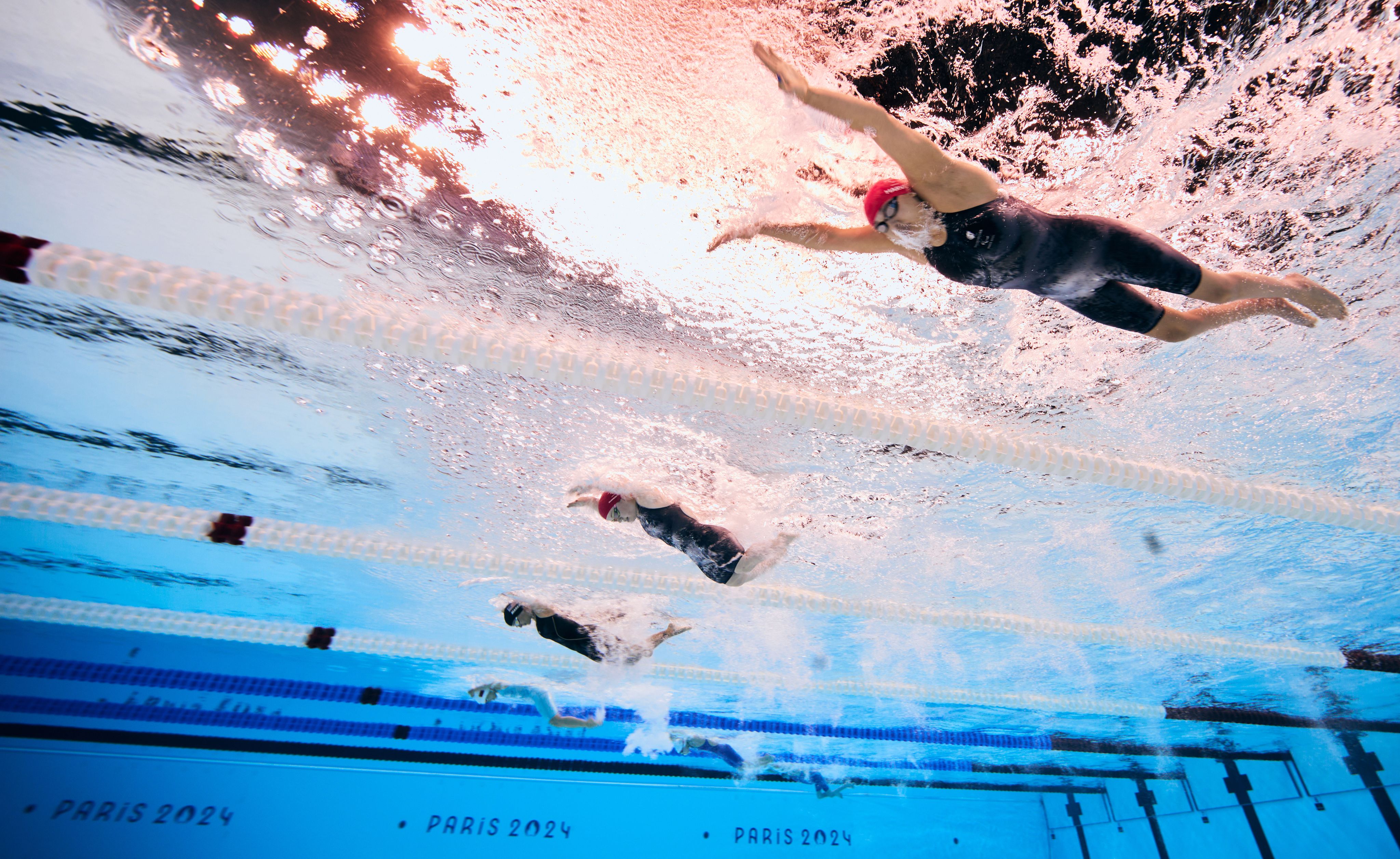 Swimmers in the pool, the camera is underwater and looking up as they swim in their individual lanes.