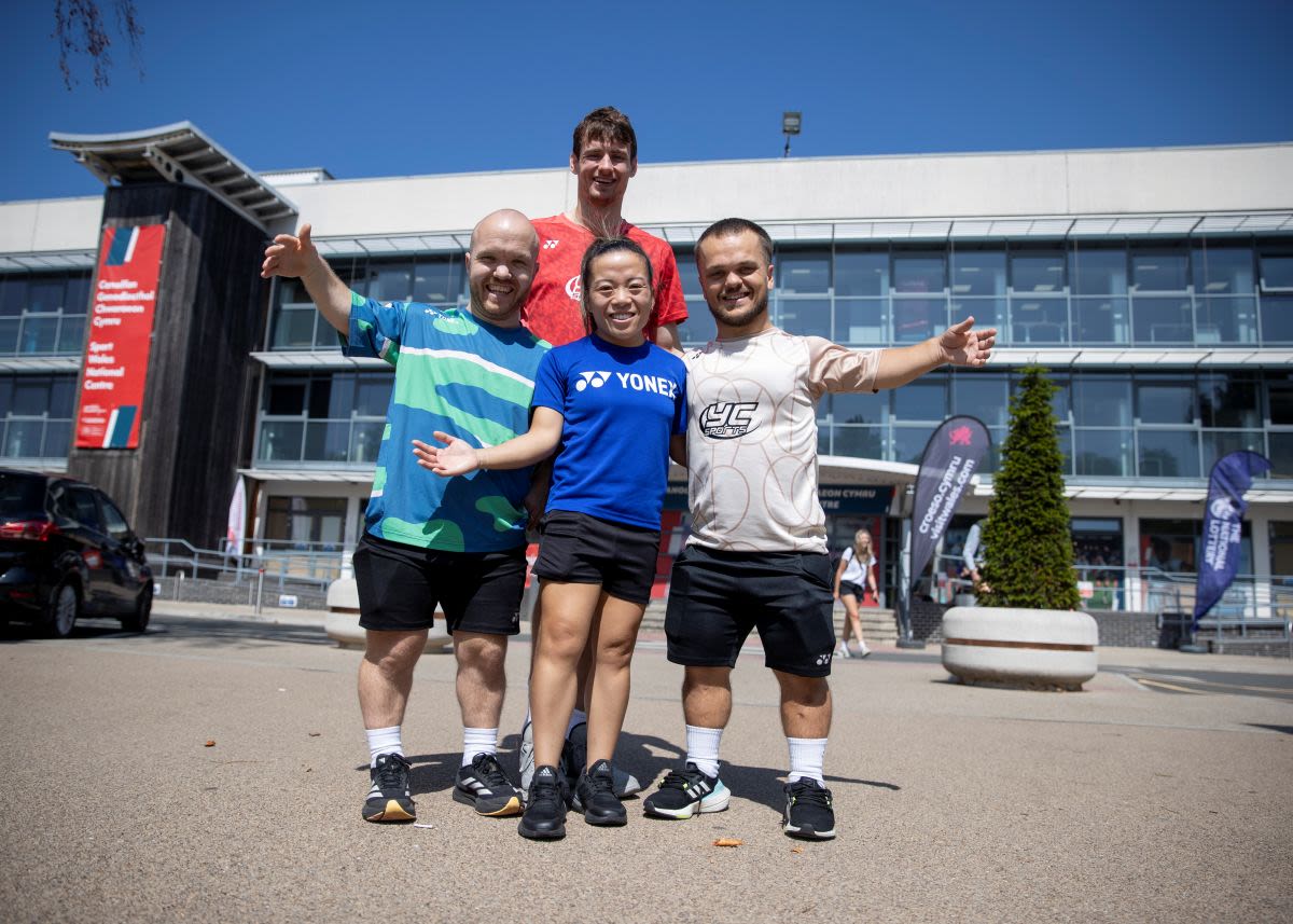 Krysten Coombs, Daniel Bethell, Rachel Choong, and Jack Shephard of Team England stand together outside the Sport Wales National Centre in Cardiff during the British & Irish Para Badminton International. They are wearing sports attire and posing with arms outstretched in a celebratory gesture. The modern building behind them has large glass windows, banners, and flags, and the scene is set under a clear blue sky.