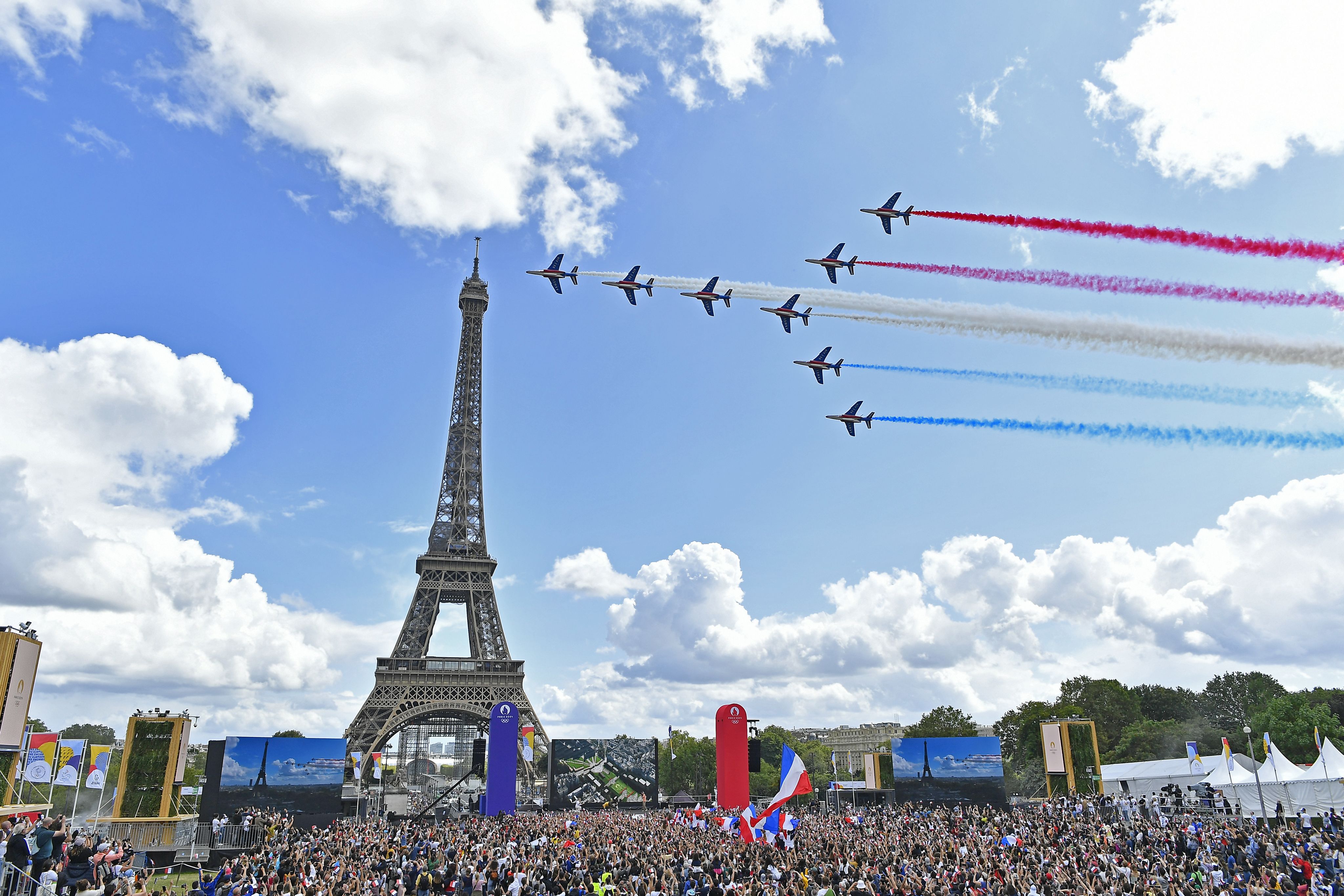 Seven acrobatic aeroplanes of the Patrouille de France fly past the Eiffel Tower in Paris, with red, white, and blue smoke trailing behind them to form a French tricolore flag.