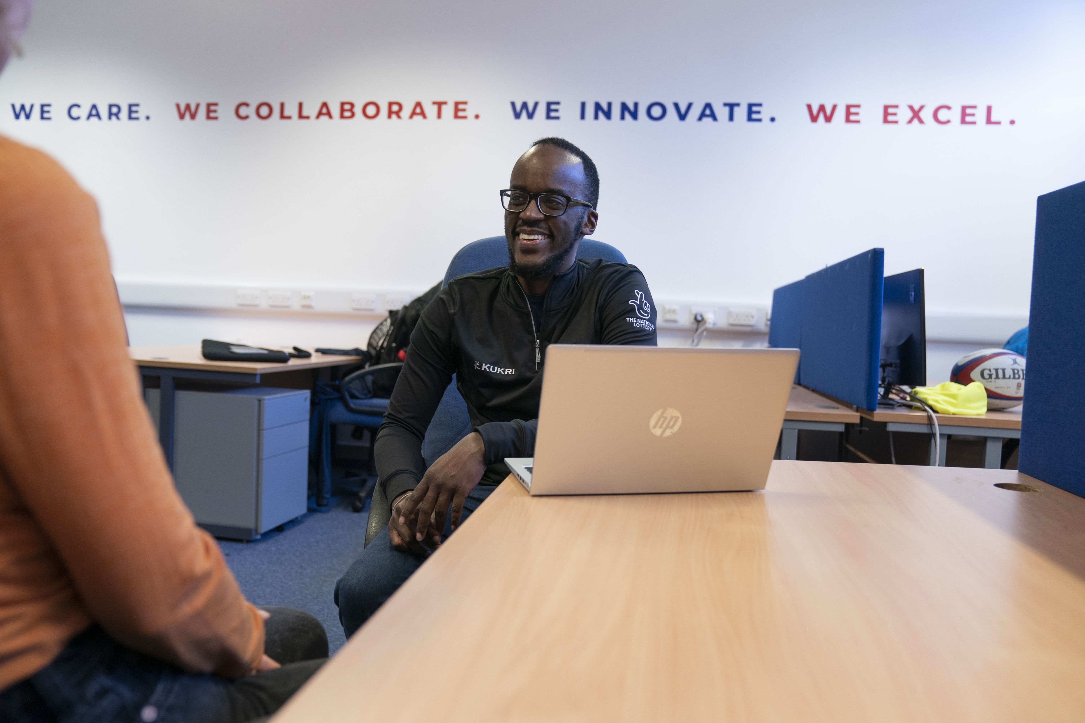 A smiling UKSI practitioner in front of a wall graphic stating 'We Care. We Collaborate. We Innovate. We Excel.'